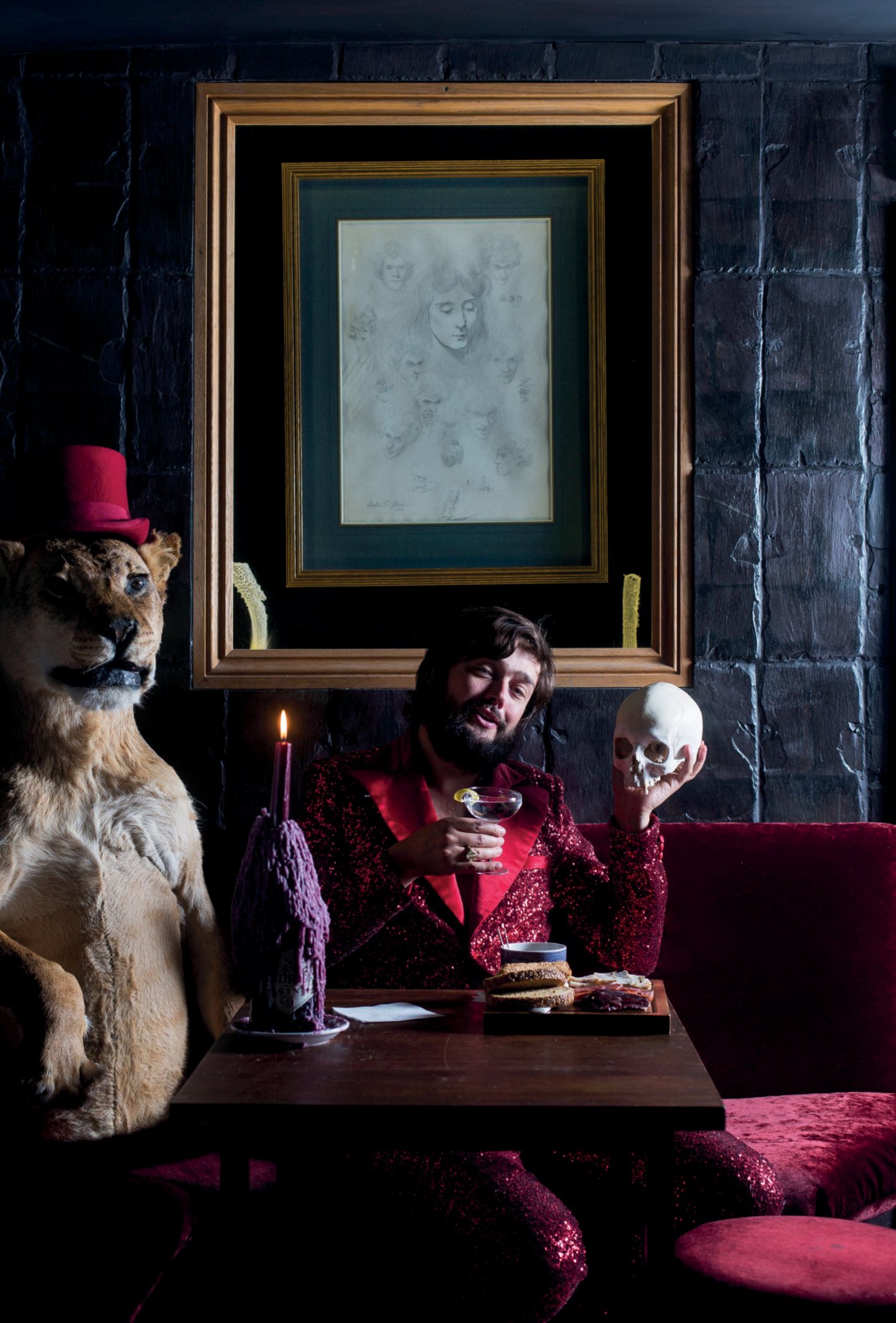 Schön schräg: Museum of Curiosities Viktor Wynd im Samtjacket sitzt auf einem Sofa, in der Hand ein Totenschädel. Daneben steht ein ausgestopfter Löwe.