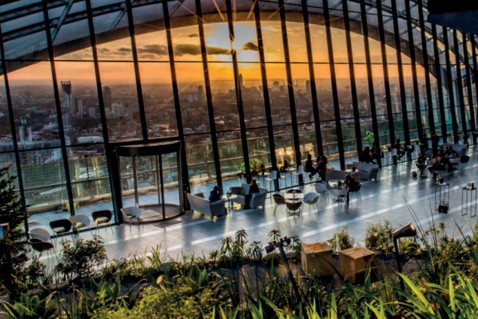 Großes, bodentiefes, halbrundes Fenster überspannt einen Platz mit Tischen und Pflanzen. Man sieht die Skyline Londons  0.6771