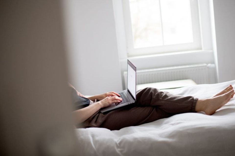 Eine Person liegt mit Laptop und Jogginghose auf einem Bett. Das Gesicht ist nicht erkennbar