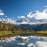 Seeufer mit Wiesen und ein paar Bäumen, verschneite Berge, die sich natürlich auch im Wasser spiegeln.