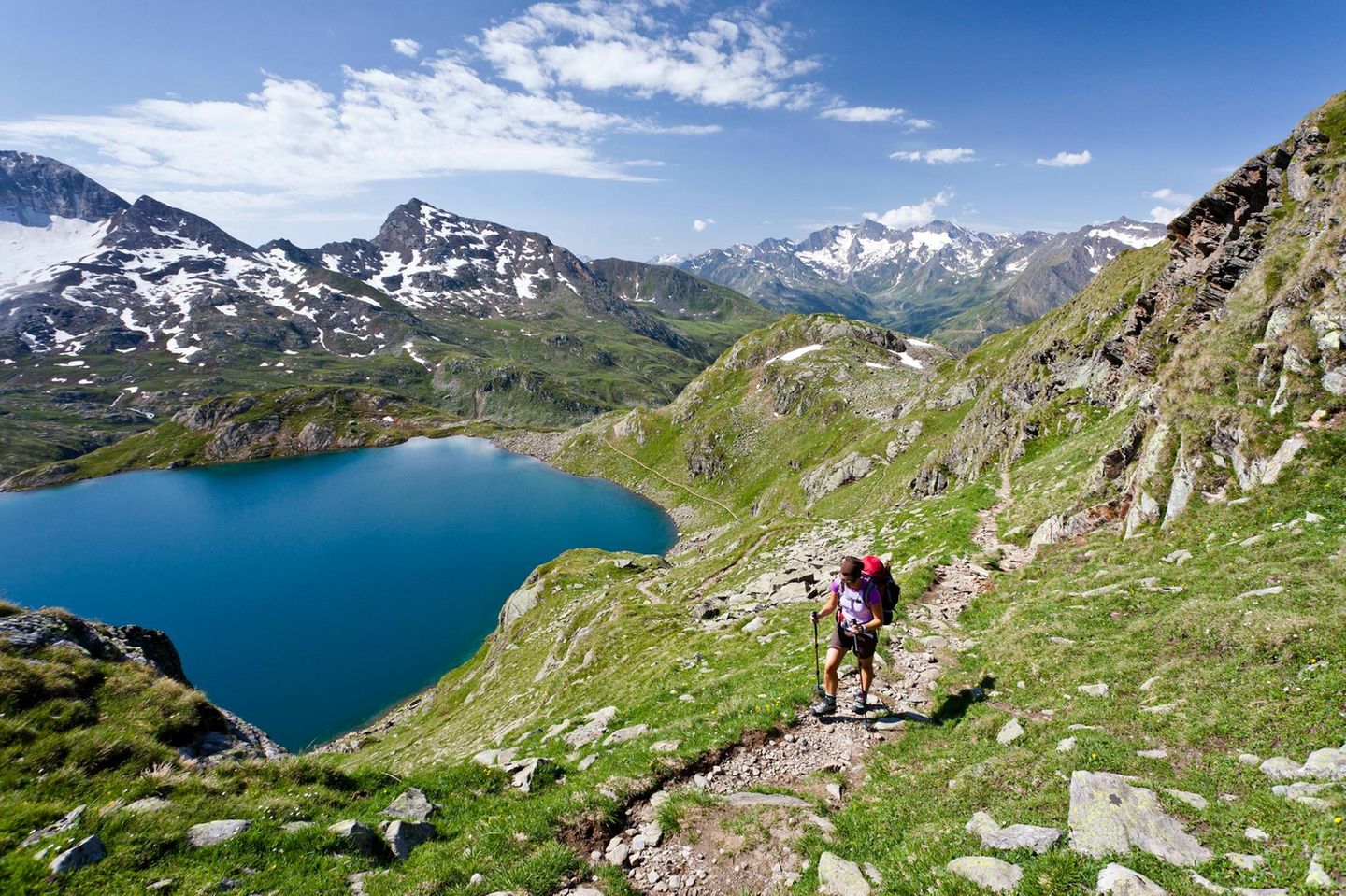 Wanderer auf Weg, der See liegt unter ihm in einer Senke, Berggipfel mit Schneeresten.