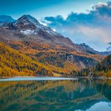 Herbstliche Lärchenwälder glühen an den Hängen, Gipfel mit Schnee, unten das stille Wasser eines großen Sees.