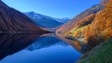 Herbstlicher Wald mit Almen, weiter hinten Schneegipfel, der See liegt unten.