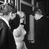 Marilyn Monroe in einem hautengen Kleid, Kennedy und andere stehen vor einer Bücherwand zusammen; sw-Foto