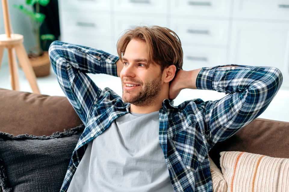 Junger Mann sitzt lächelnd mit hinter dem Kopf verschränkten Armen auf dem Sofa  1.5026