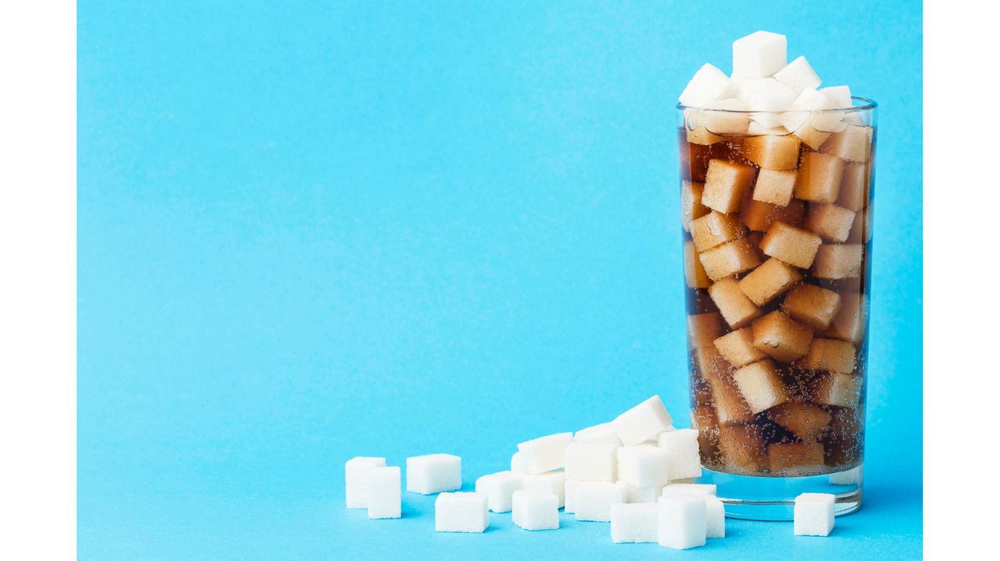 Trinkglas randvoll gefüllt mit Cola und Zuckerwürfeln. Ein Teil der Zuckerwürfel liegt neben dem Glas.