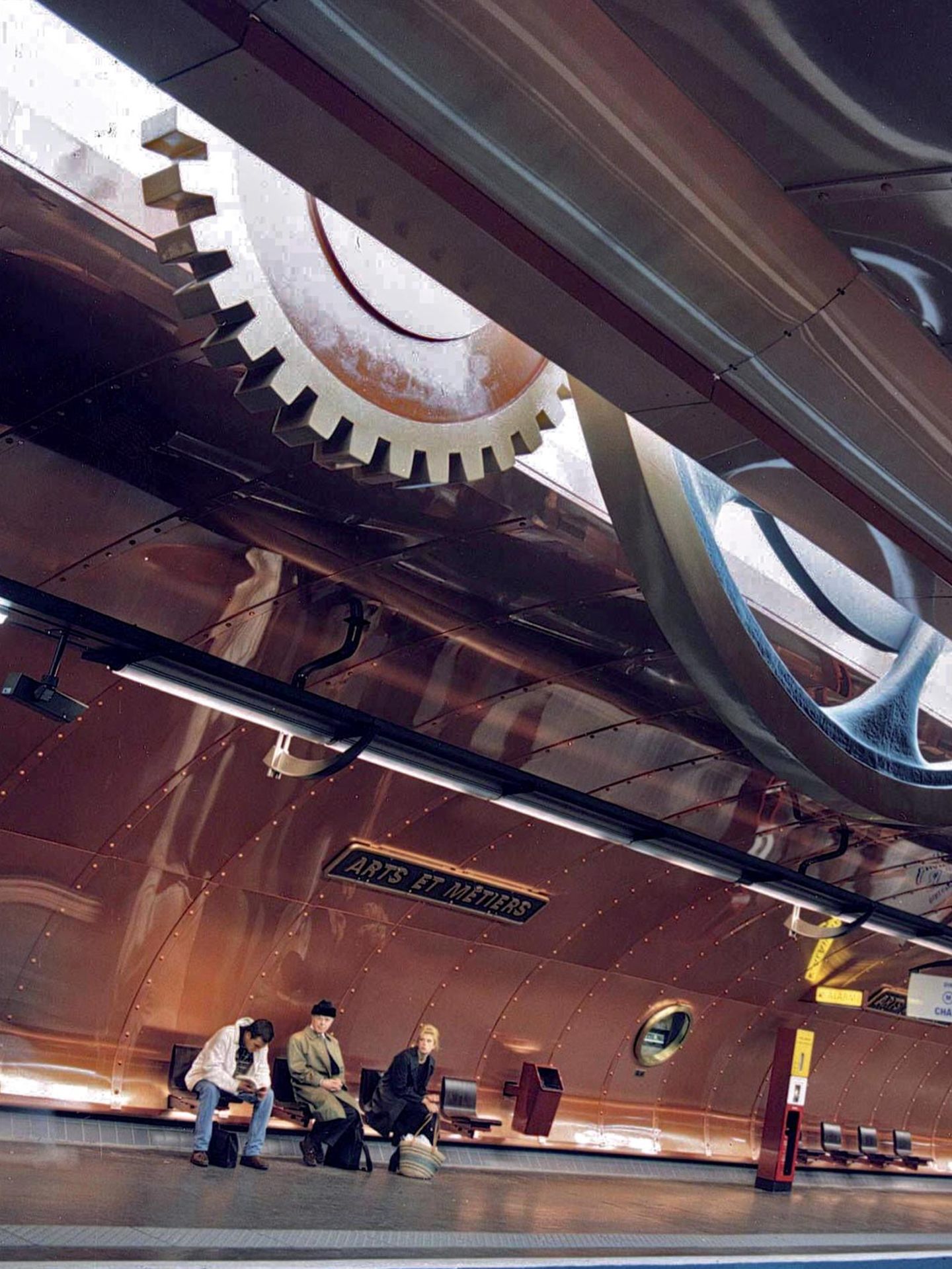 Wartebank mit Menschen, die kupfernen Wände glänzen, zwei Zahnräder schieben sich durchs Dach.