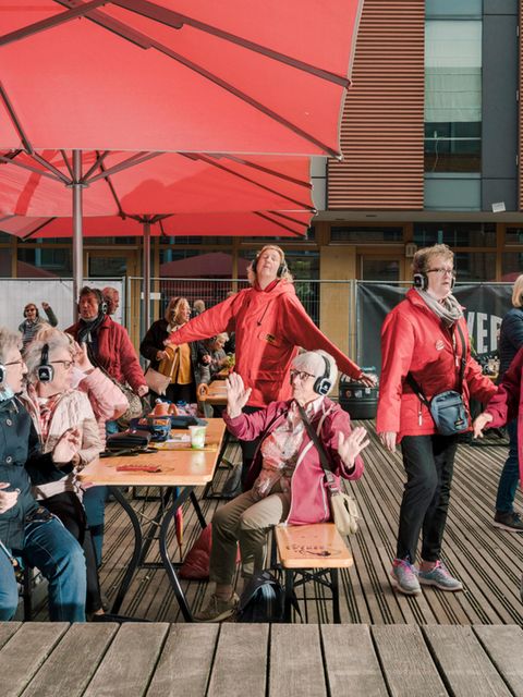5 ältere Frauen mit Kopfhörern auf den Ohren sitzen und stehen draussen in einem Outdoorcafe, tanzen und haben Spass miteinander