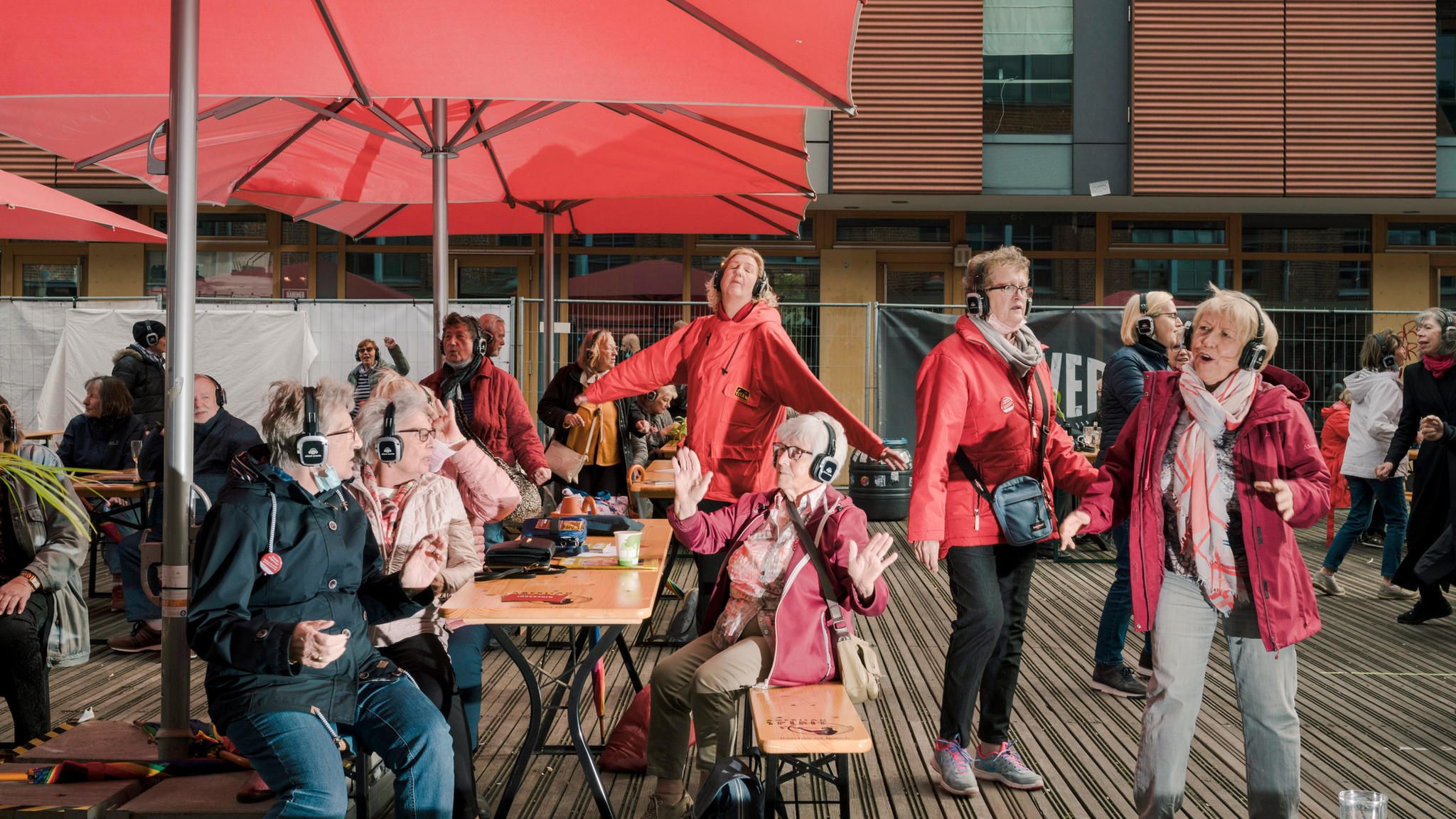 5 ältere Frauen mit Kopfhörern auf den Ohren sitzen und stehen draussen in einem Outdoorcafe, tanzen und haben Spass miteinander