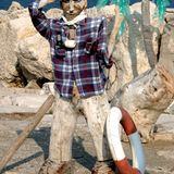 Helle Felsen, dahinter das Meer, im Vordergrund Figur aus Treibholz mit Kapitänsmütze, die salutiert.