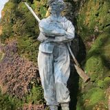 Mit Moos bewachsener Felsen, mannsgroße Statue mit Gewehr, Kappe und verschränkten Armen