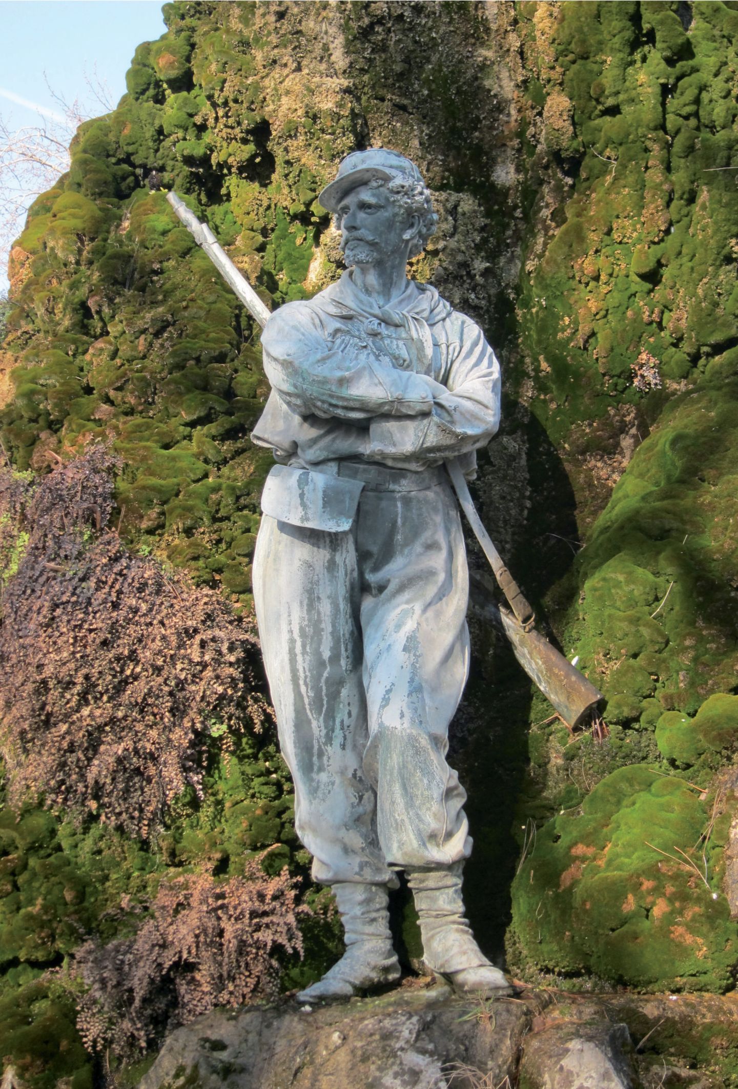 Mit Moos bewachsener Felsen, mannsgroße Statue mit Gewehr, Kappe und verschränkten Armen
