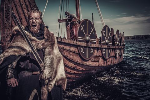 Ein bärtiger Wikinger mit Fellumhang steht vor einem Schiff: schreiend