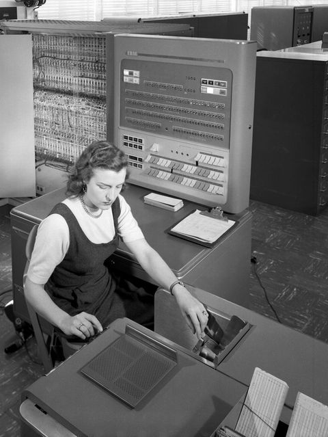 Frau arbeitet am Schreibtisch während ein Mann hinter ihr etwas an einem technischen Gerät überprüft