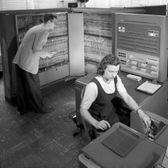 Frau arbeitet am Schreibtisch während ein Mann hinter ihr etwas an einem technischen Gerät überprüft