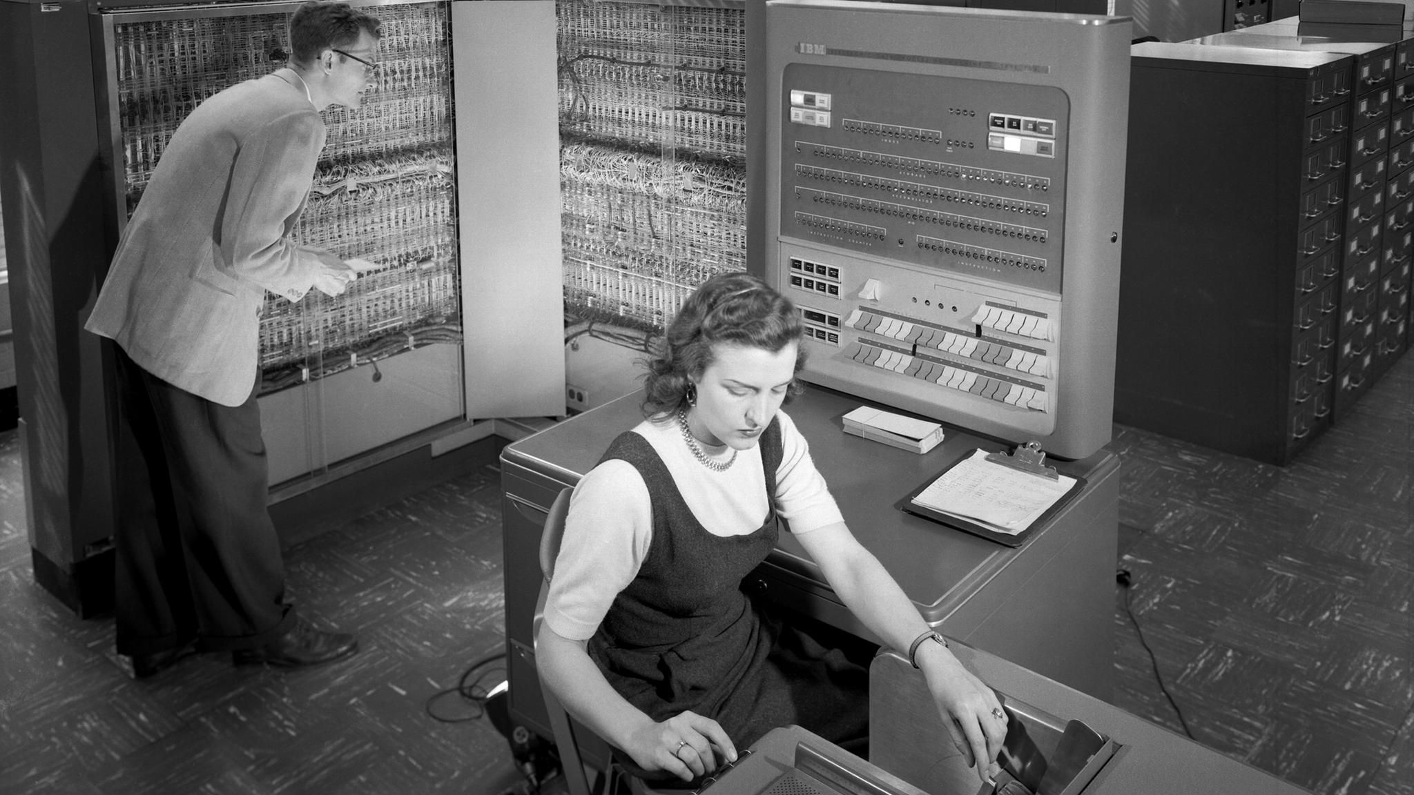 Frau arbeitet am Schreibtisch während ein Mann hinter ihr etwas an einem technischen Gerät überprüft Frau arbeitet am Schreibtisch während ein Mann hinter ihr etwas an einem technischen Gerät überprüft