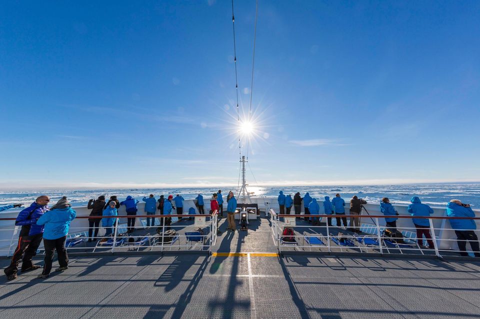 Viele Menschen in blauen Jacken stehen bei Sonnenschein und kaltem Wetter an der Reling eines Schiffes
