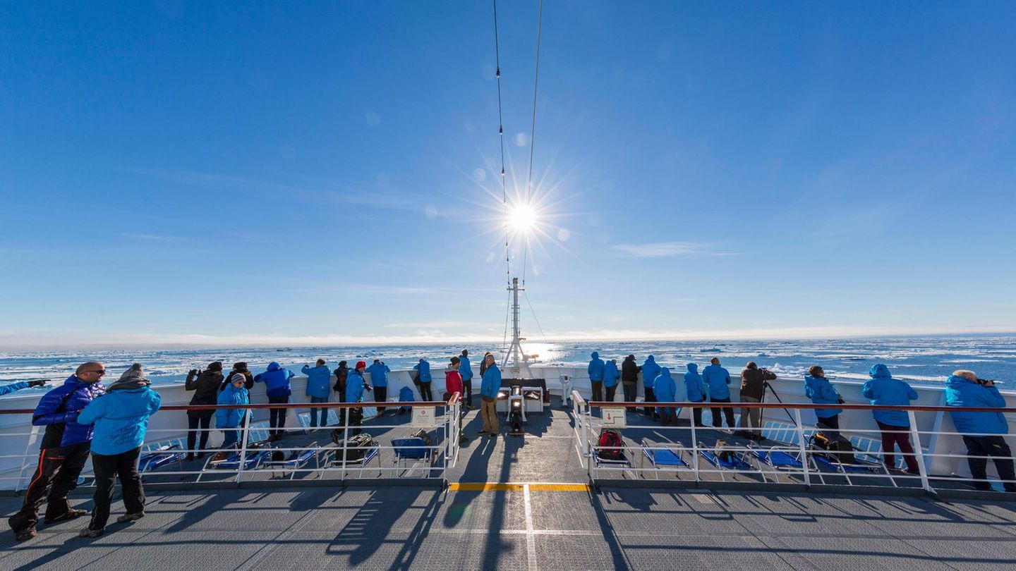 Viele Menschen in blauen Jacken stehen bei Sonnenschein und kaltem Wetter an der Reling eines Schiffes