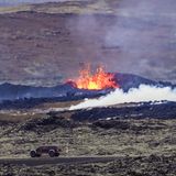 Ein Auto fährt auf einer Straße auf Island, dahinter schießt eine Lavafontäne aus dem Boden
