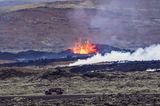 Ein Auto fährt auf einer Straße auf Island, dahinter schießt eine Lavafontäne aus dem Boden