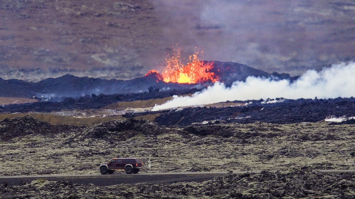 Ein Auto fährt auf einer Straße auf Island, dahinter schießt eine Lavafontäne aus dem Boden