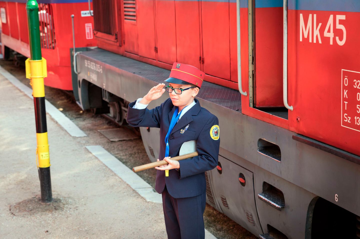 Vor einer Diesellok steht ein Junge mit Kelle in Bahnuniform und salutiert