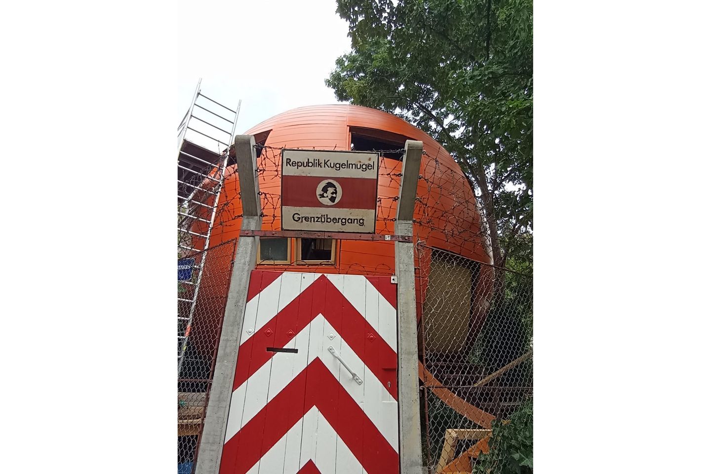 Tür mit Stacheldraht, der Grenzübergang zur Mikronation, eine große runde Kugel, daneben Bäume