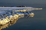 felsiger, schneebedeckter Strandabschnitt am Meer, in der Ferne sind Häuser zu sehen