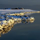 felsiger, schneebedeckter Strandabschnitt am Meer, in der Ferne sind Häuser zu sehen