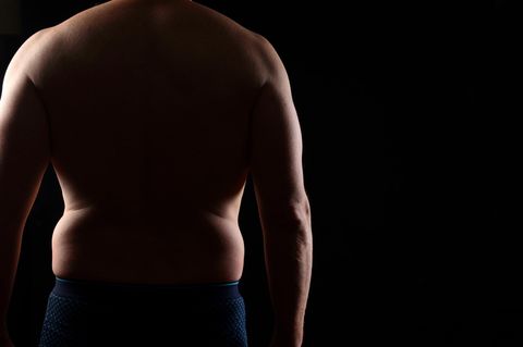 übergewichtiger Mann mit freiem Oberkörper von hinten vor dunklem Hintergrund