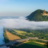 Blick über einen Fluss, daneben ein bewaldeter Berg mit tempelartigem Bau darauf, Nebelschwade