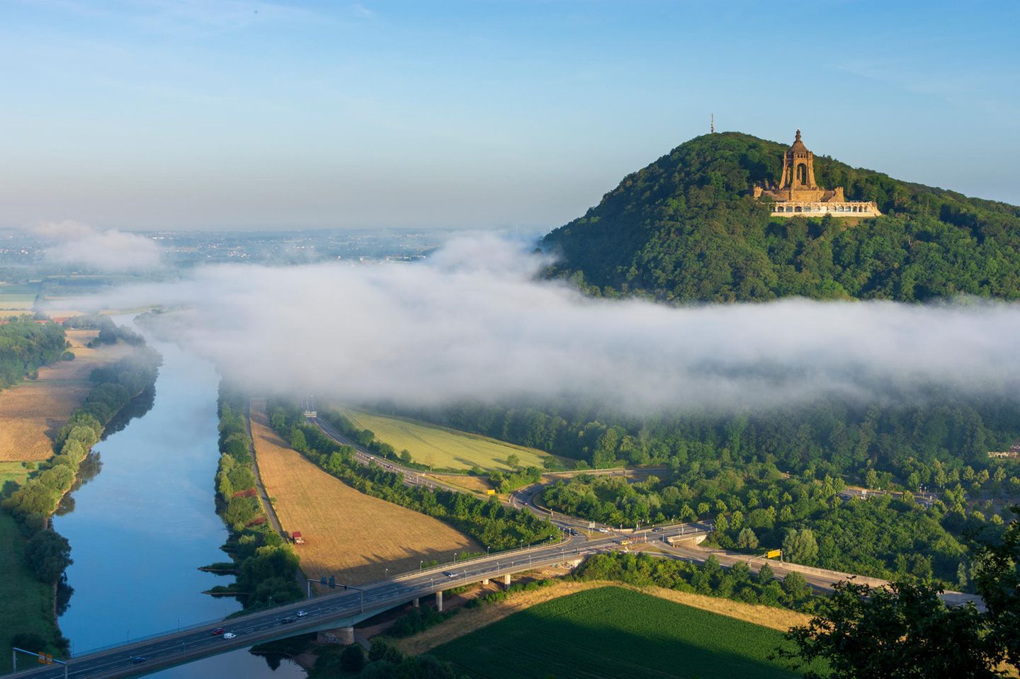 Blick über einen Fluss, daneben ein bewaldeter Berg mit tempelartigem Bau darauf, Nebelschwade