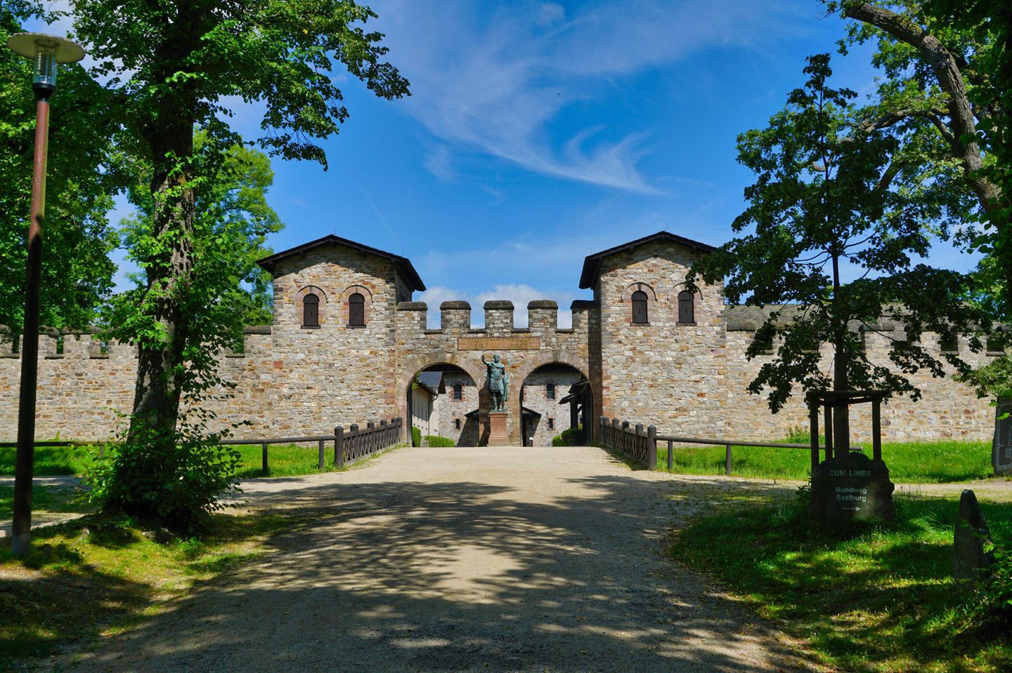 Steinbau gesäumt von Bäumen mit zwei Eingangstoren, dazwischen eine römische STatue