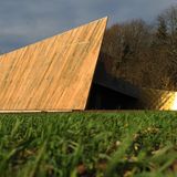 Gebäude aus Holz mit spitzer Form, dahinter Bäume