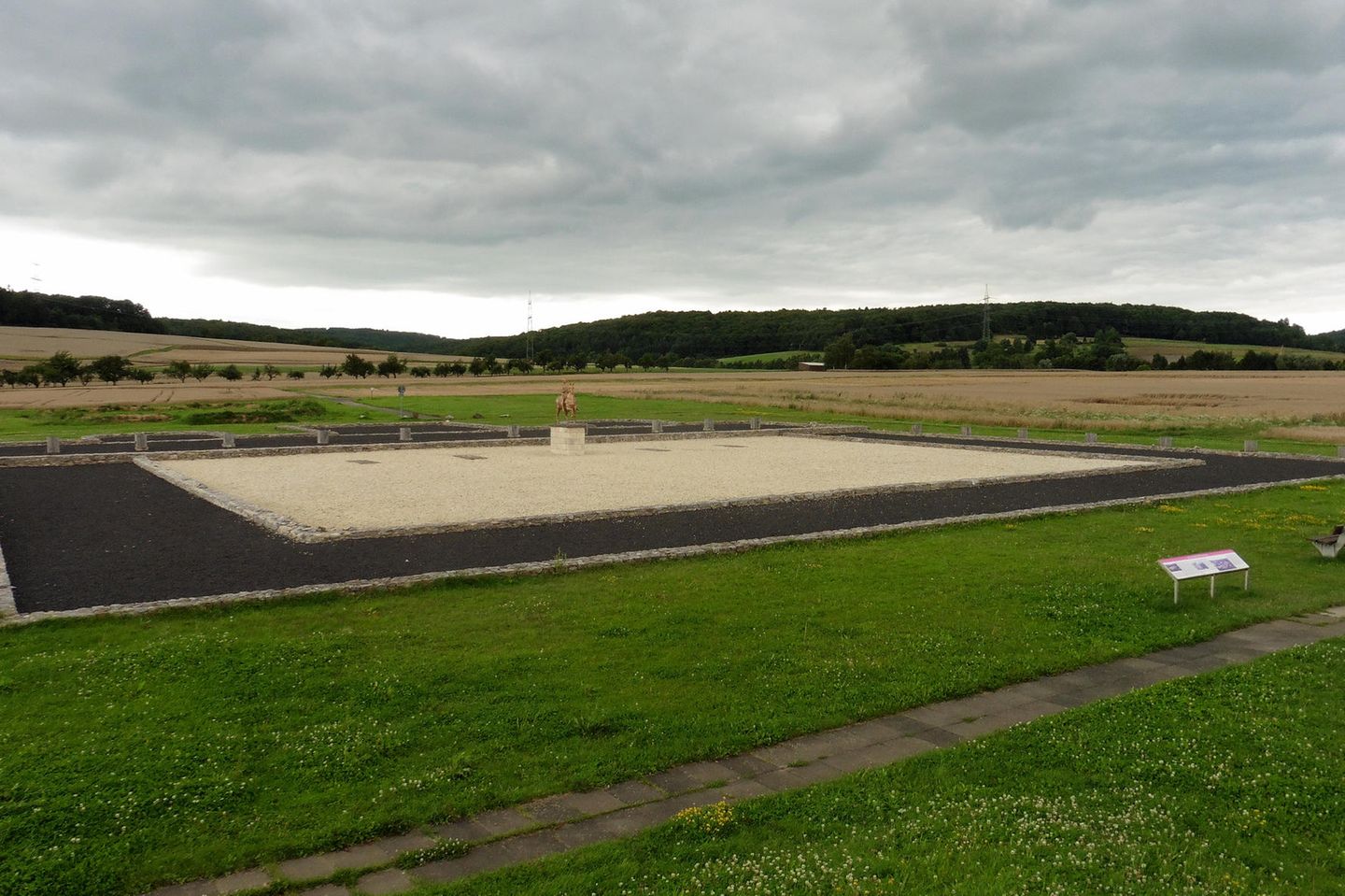 Feld und Wiese, in der Mitte befindet sich ein Quadrat, dessen Mitte eine Statue steht