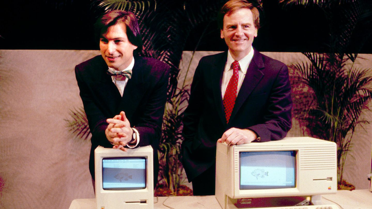 Steve Jobs lehnt auf dem Macintosh PC, daneben John Sculley hinter einem anderen Modell