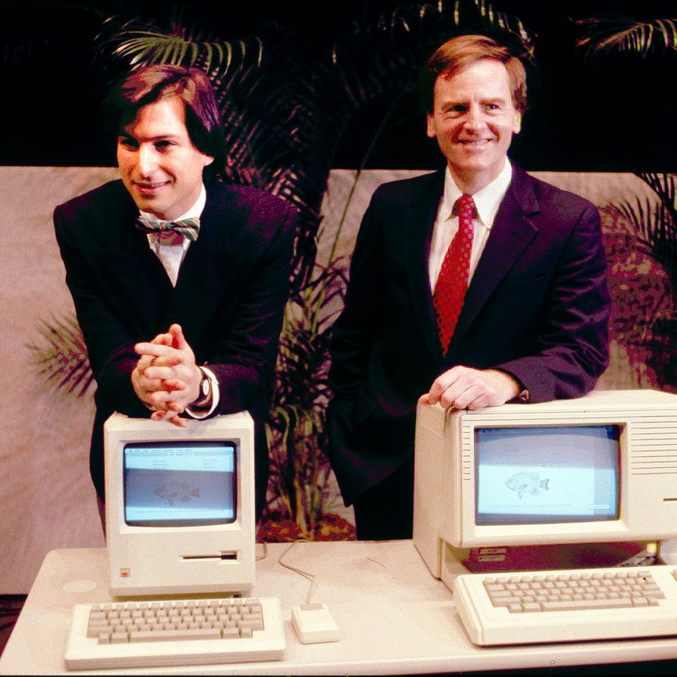Steve Jobs lehnt auf dem Macintosh PC, daneben John Sculley hinter einem anderen Modell