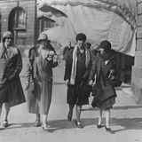 vier Frauen mit Hut spazieren vor Häusern