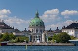 Kirche mit grünem Dach umgeben von Häusern, davor Treppen am Fluss