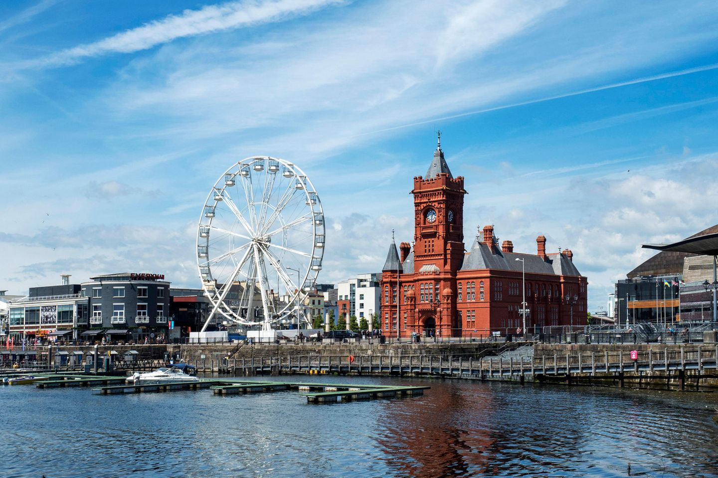 Großes rotes Gebäude, Riesenrad und Wohnhäuser vor einem Fluss mit Steg
