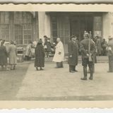 Mehrere Menschen vor Gebäude mit großen Fenstern, darunter ein Mann mit Gewehr und ein Mann in weißem Kittel