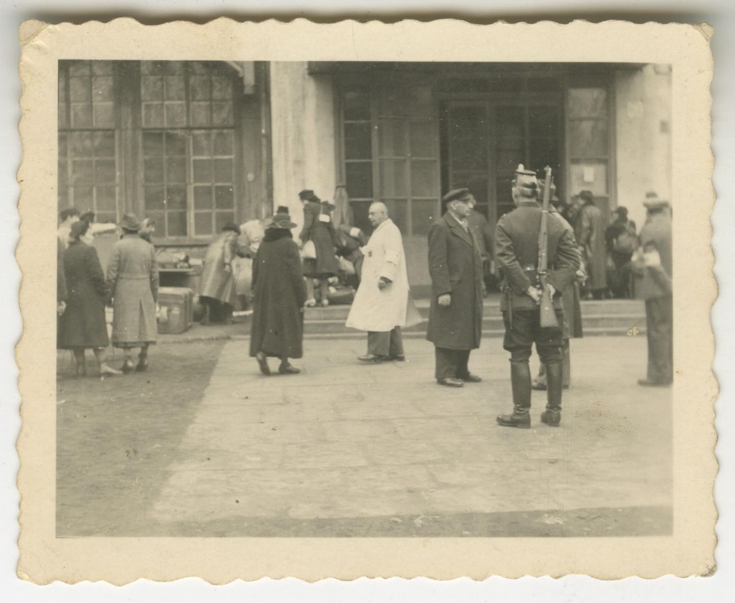 Unter dem Kommando der Gestapo Mehrere Menschen vor Gebäude mit großen Fenstern, darunter ein Mann mit Gewehr und ein Mann in weißem Kittel