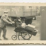 Mann schiebt Frau im Rollstuhl, Lastwagen im Hintergrund