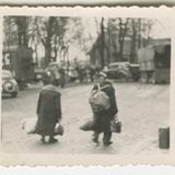 Zwei Personen mit Rucksack und Koffer laufen auf Lastwagen und Autos zu