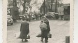Zwei Personen mit Rucksack und Koffer laufen auf Lastwagen und Autos zu
