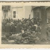 Menschenmenge vor einem Gebäude, auf Stühlen sitzend und stehend, an einem Baum daneben ist ein Absperrungsseil gespannt