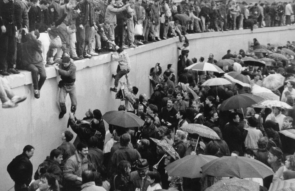 Menschen stehen auf der Mauer, unten eine Menschenmenge mit Regenschirmen, vereinzelt wird versucht, die Mauer hochzuklettern
