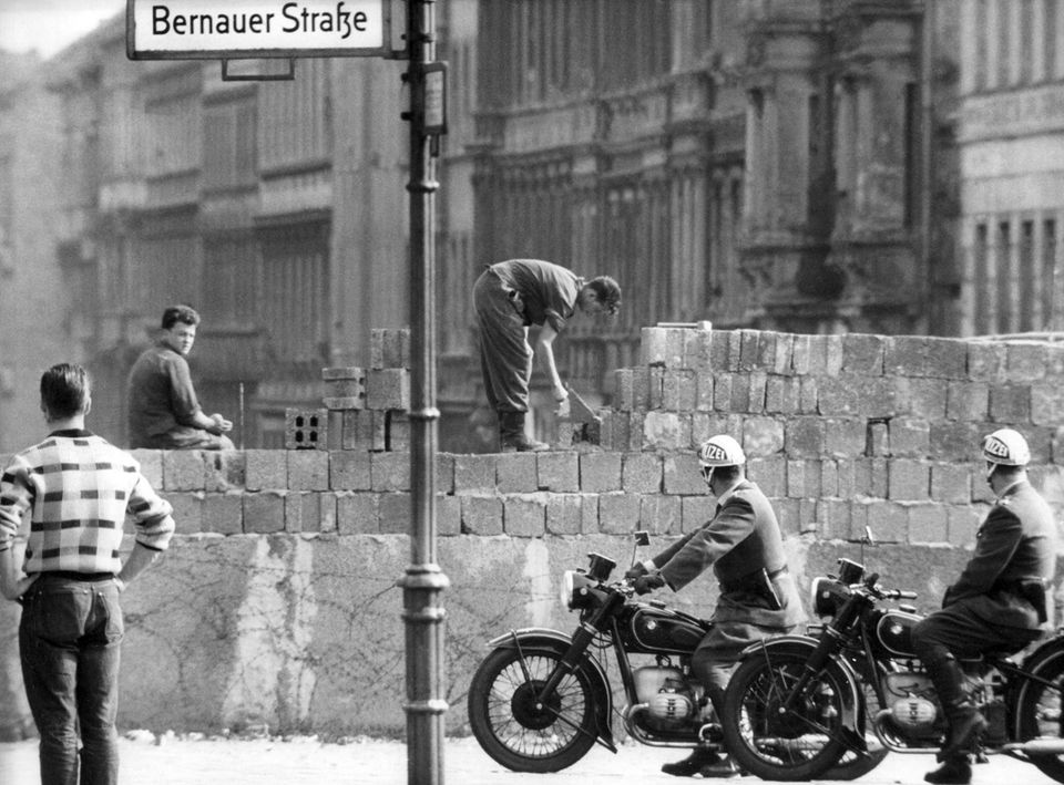 Straßenschild mit der Aufschrift "Breslauer Straße", dahinter Arbeiter an Mauer, die von Polizist auf Motorrad beobachtet werden