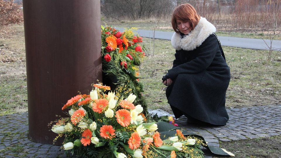 Karin Gueffroy hockt vor Blumenkränzen an Gedenkort