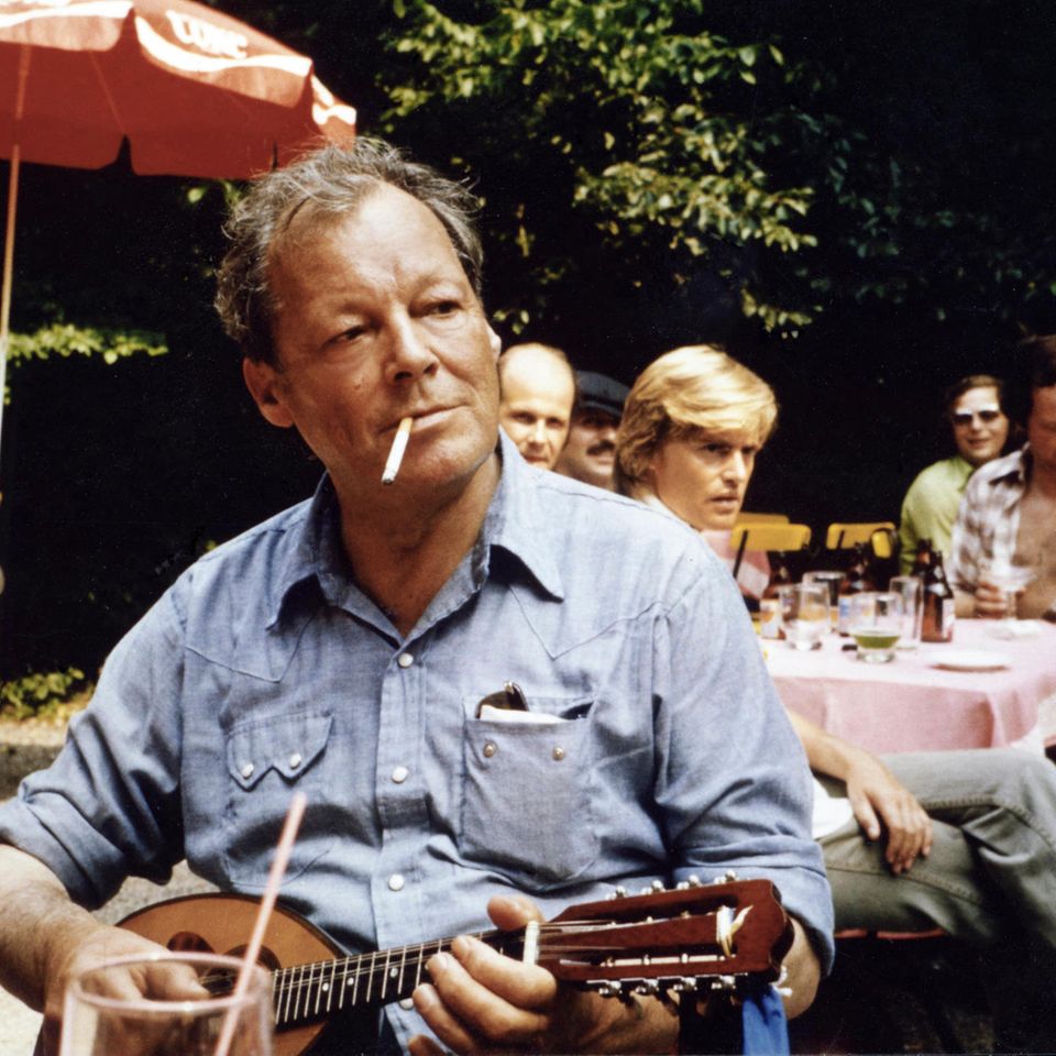Willy Brandt sitzt und spielt Mandoline mit Zigarette im Mund, im Hintergrund ein Sonnenschirm und Menschen an Tischen